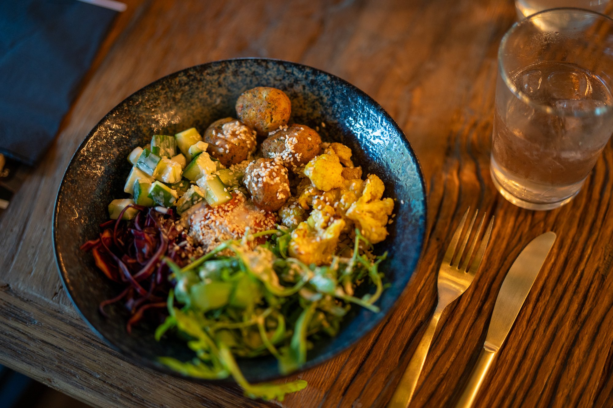 Eine Schüssel Salat auf einem Holztisch in einem Restaurant. Die Schüssel ist dunkelblau mit einer gesprenkelten Textur. Der Salat besteht aus gemischtem Grün, Rotkohl, Gurken und Falafelbällchen. Die Falafelbällchen sind goldbraun und sehen knusprig aus. Der Salat ist mit Sesamsamen und einem cremigen Dressing garniert. Neben der Schüssel stehen ein Glas Wasser und eine goldene Gabel und ein Messer. 