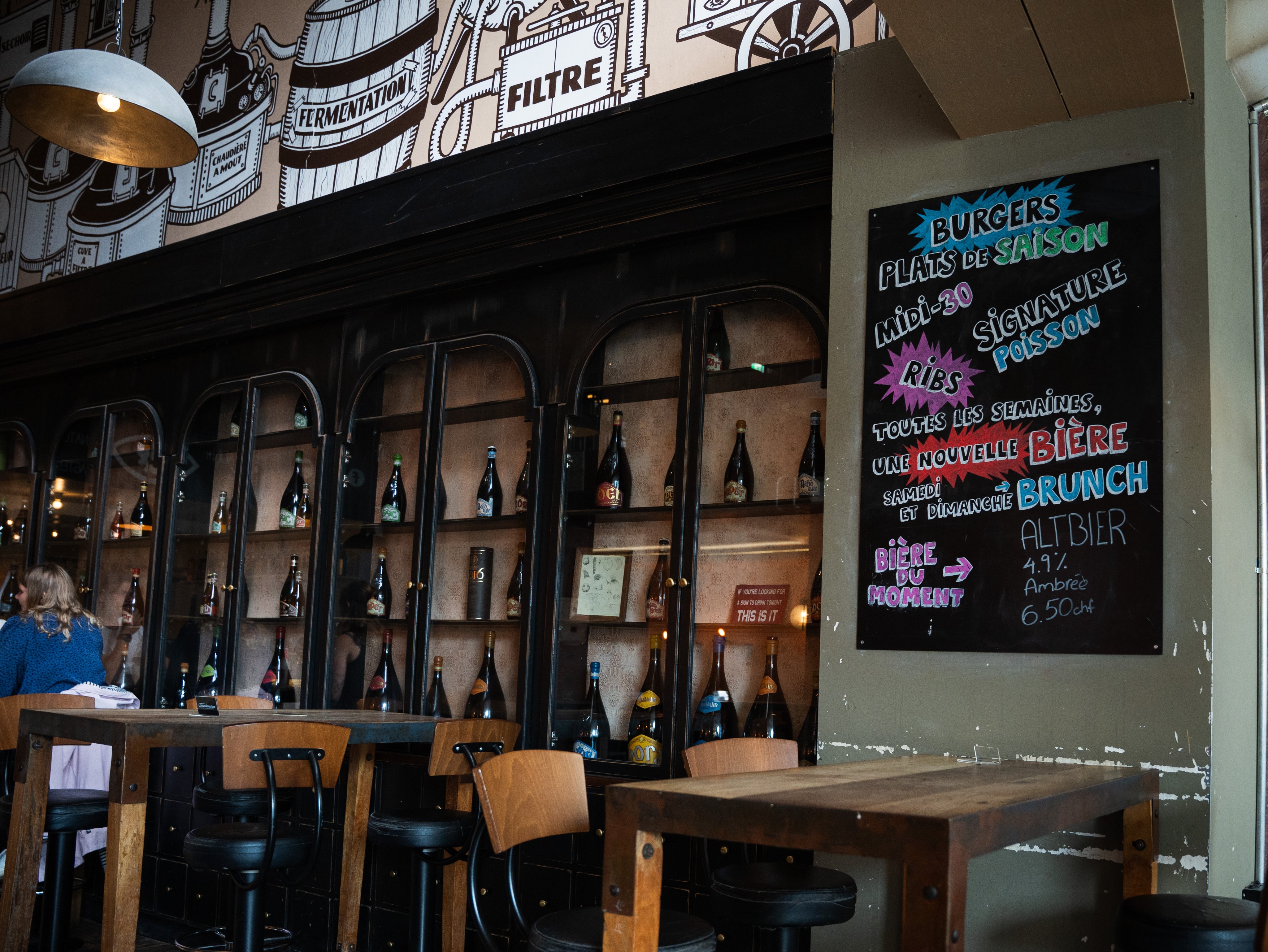 Des bouteilles sont posées sur une étagère contre le mur, devant laquelle sont installées des tables et des chaises.