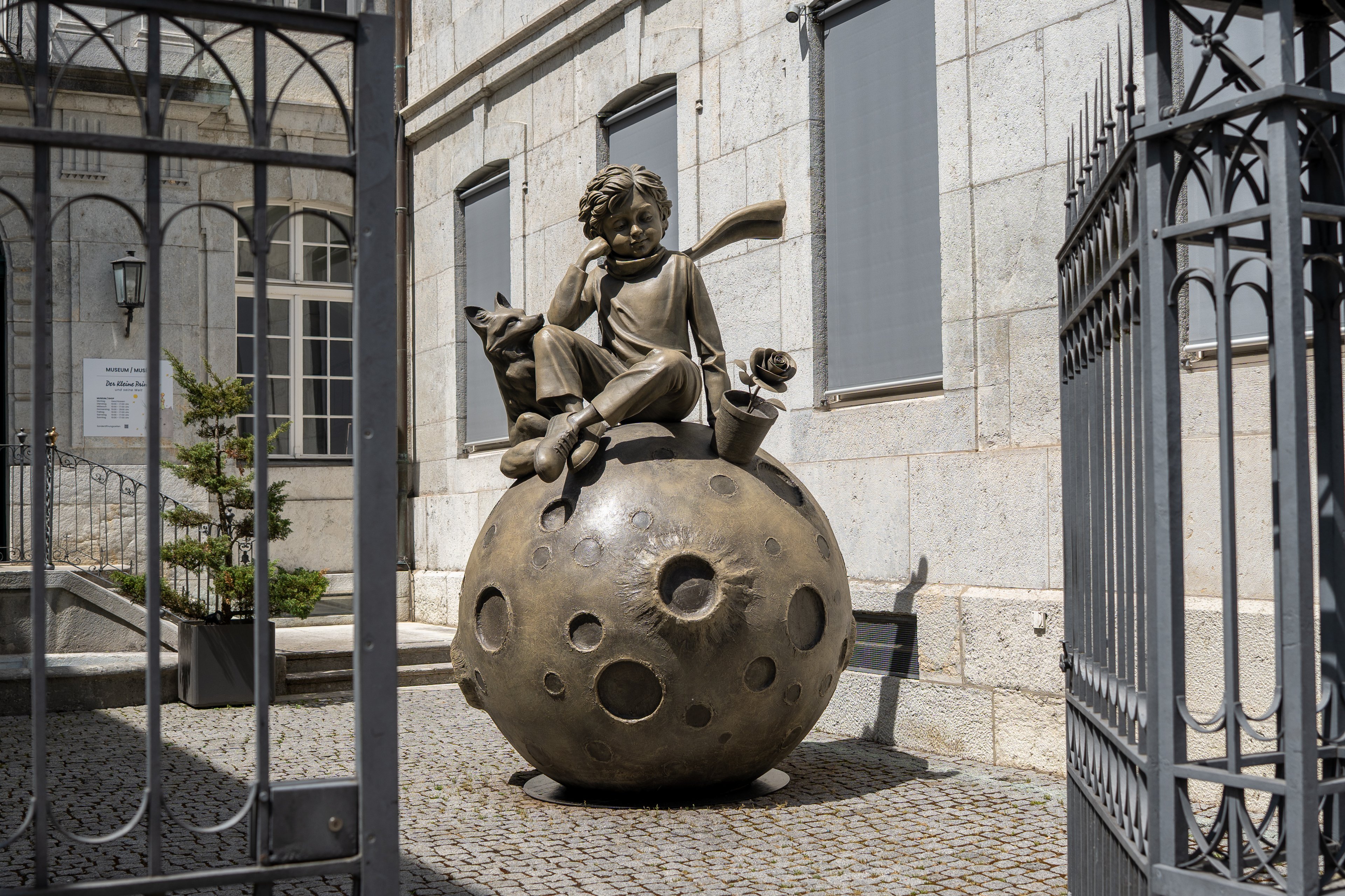 Hinter der geöffneten Türe eines Gittertors befindet sich vor einer Hausfassade ein grosses Objekt. Es zeigt eine Kugel, auf der eine Figur sitzt.