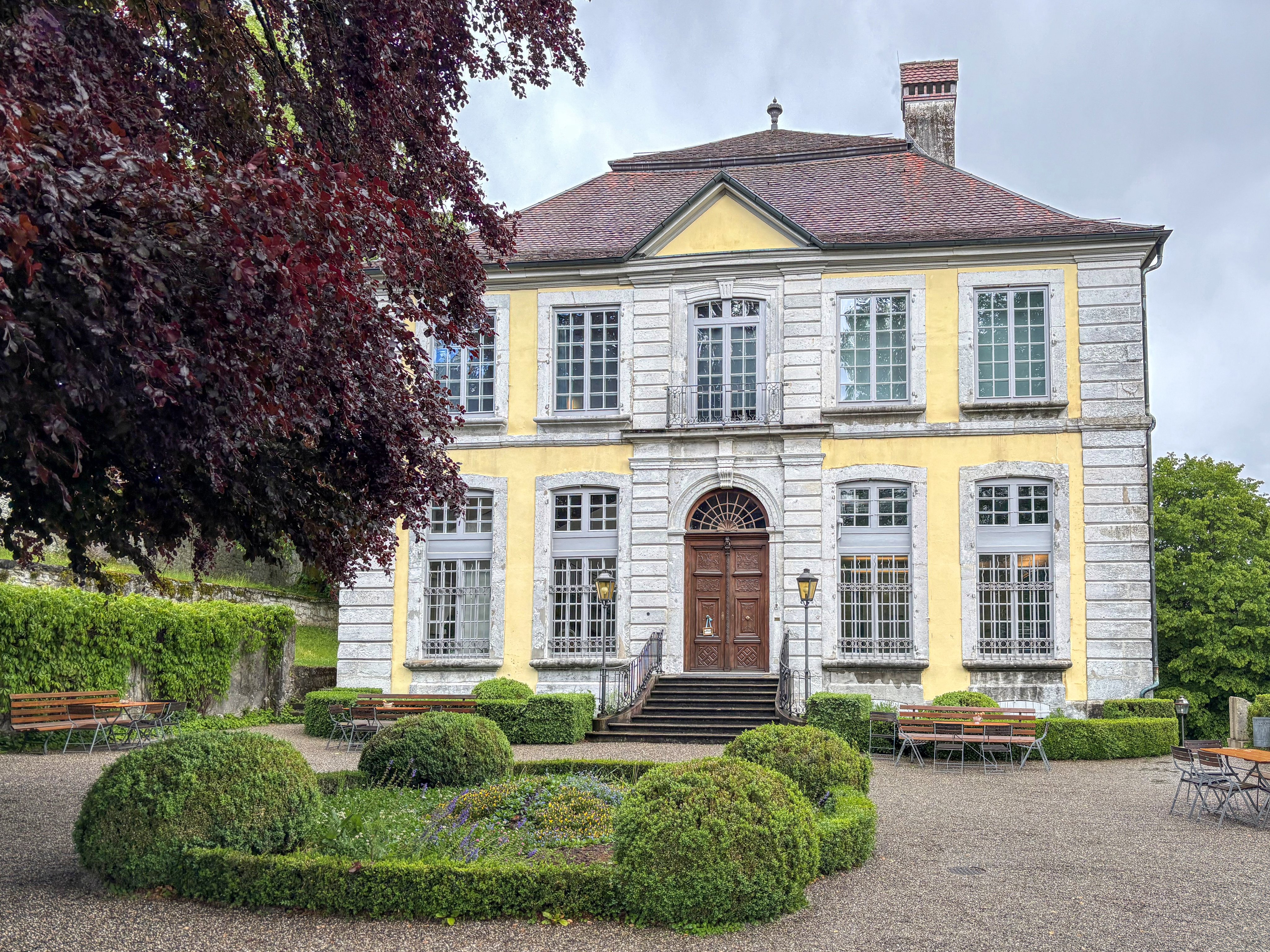 Ein altes zweistöckiges Haus mit einem Kiesplatz sowie mit akkurat gestutzten, niedrigen Hecken davor.