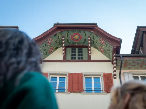 Über die Schulter einer Frau sieht man ein mit Blumen bemalter Dachgiebel.