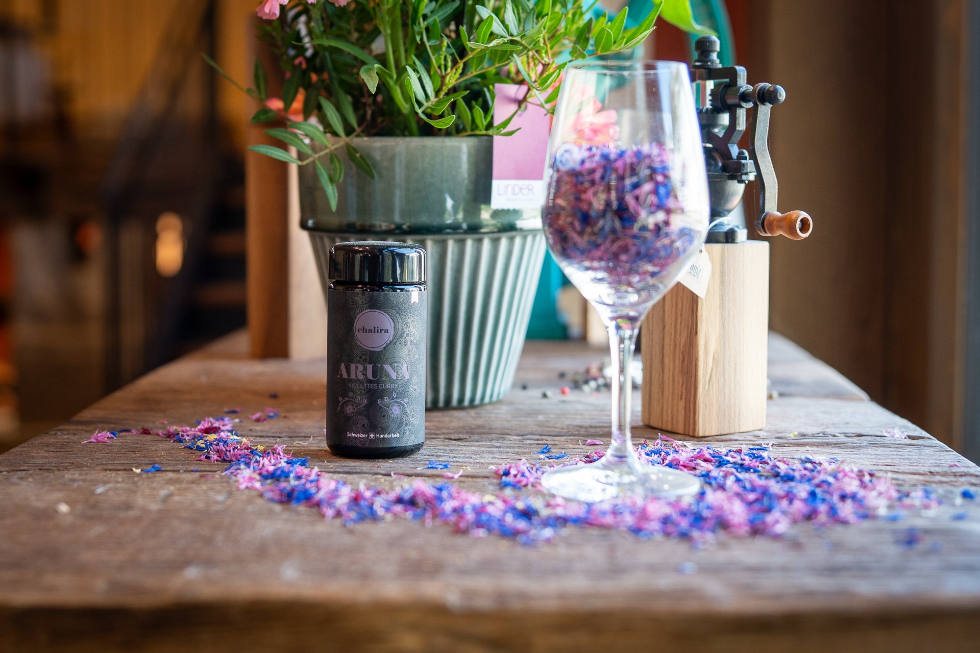 Auf einem alten Holztisch steht eine Dose mit einer Gewürzmischung. Daneben befindet sich ein Glas, das mit violetten Blüten gefüllt ist.