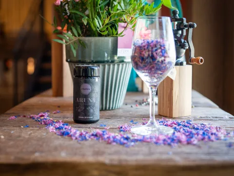 Auf einem alten Holztisch steht eine Dose mit einer Gewürzmischung. Daneben befindet sich ein Glas, das mit violetten Blüten gefüllt ist.