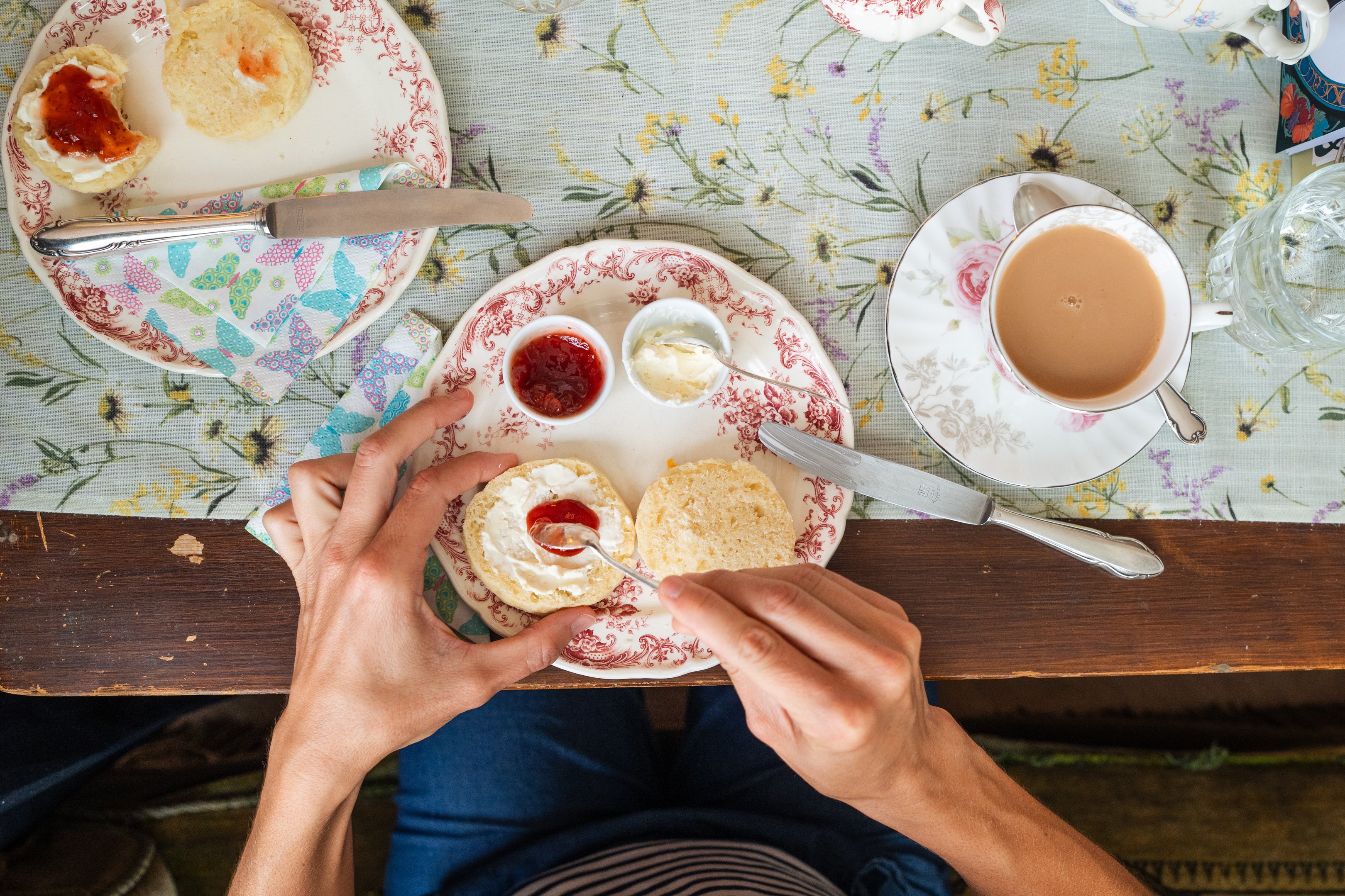 Vue d’en haut sur une assiette en porcelaine avec scones et tasse de thé. Une personne met de la confiture sur un scone.
