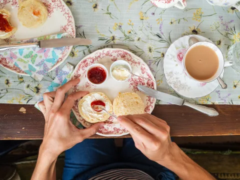 Vue d’en haut sur une assiette en porcelaine avec scones et tasse de thé. Une personne met de la confiture sur un scone.