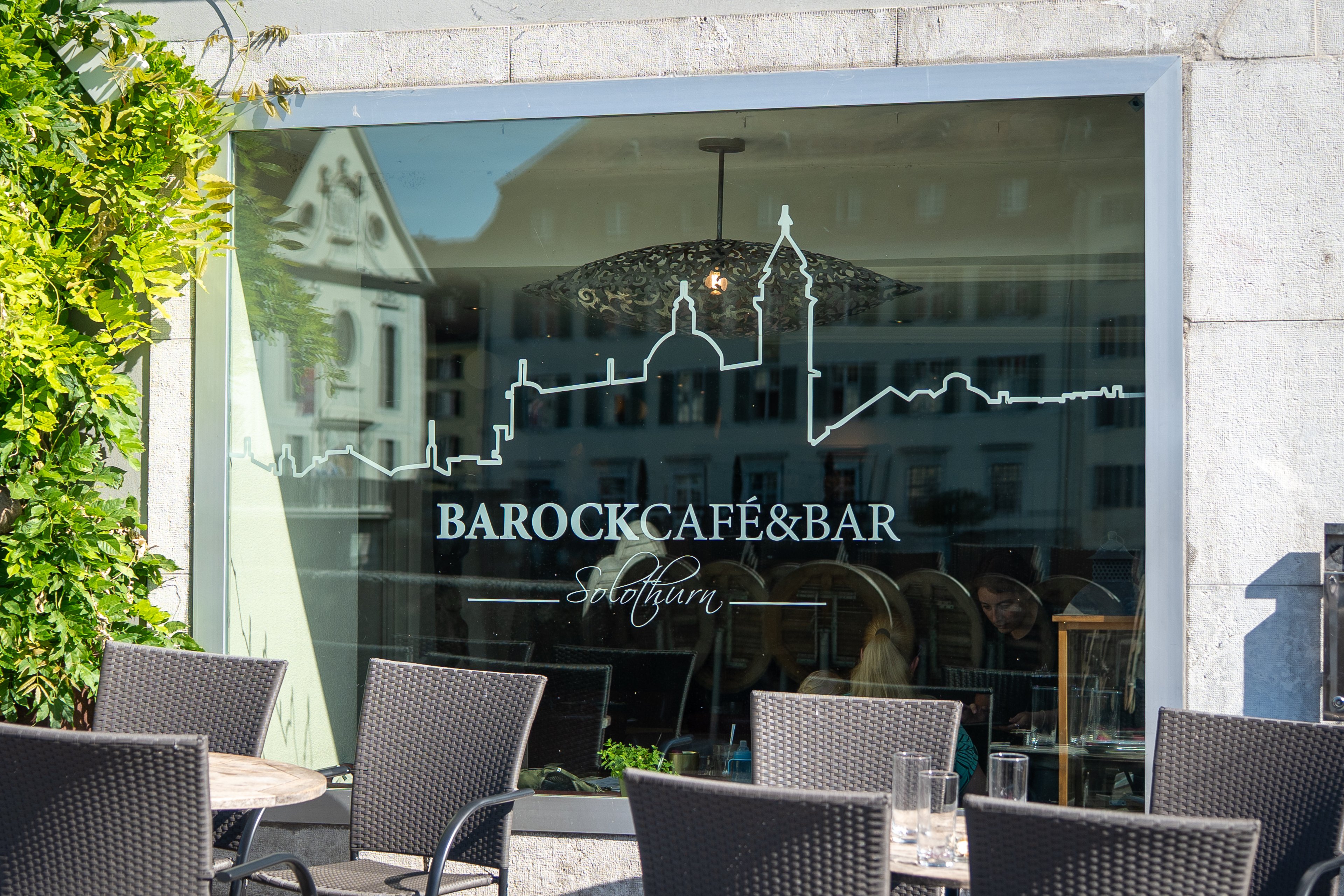 Ein Schaufenster mit der Aufschrift «Barock Café & Bar», davor stehen Tische und Stühle.
