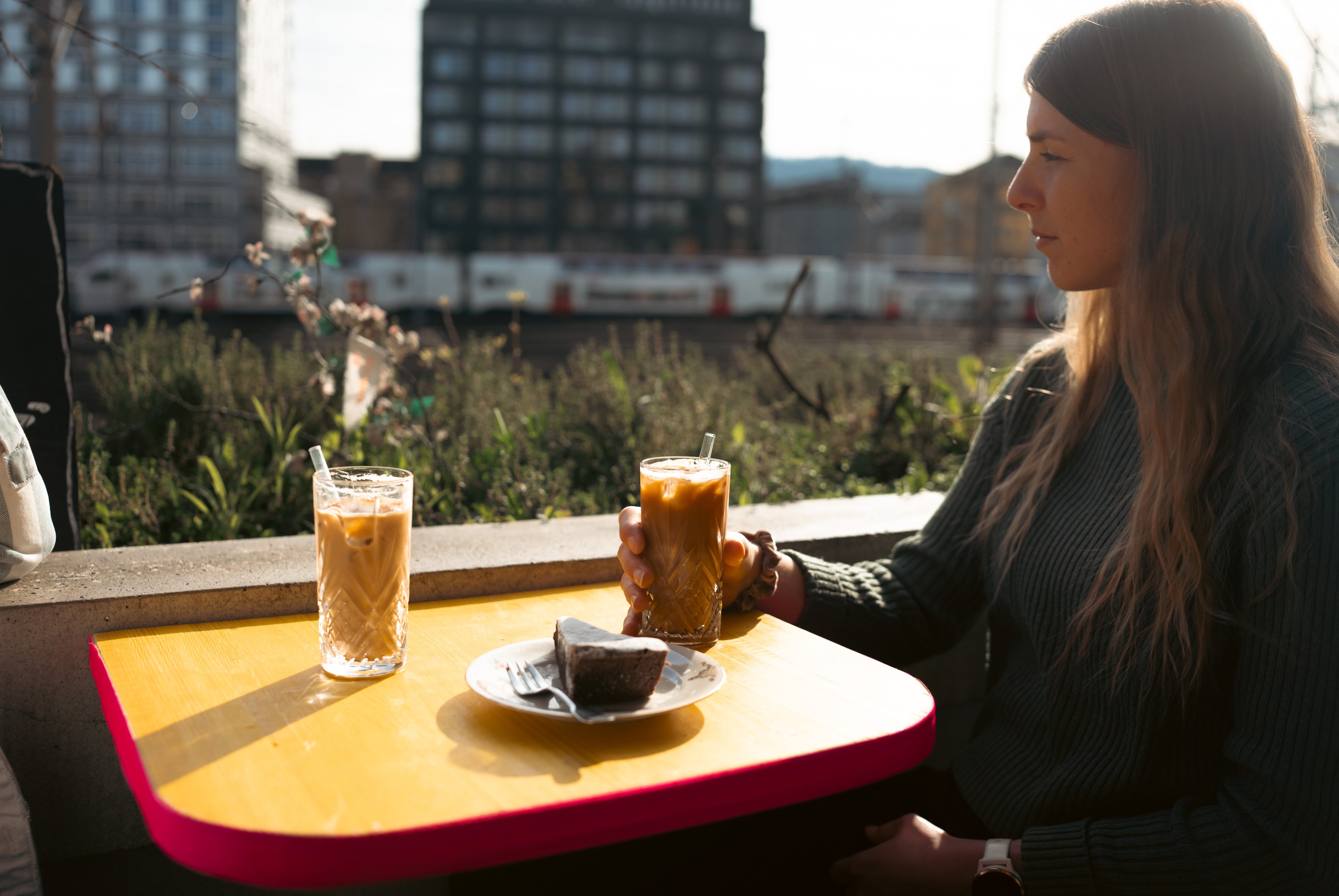 Une femme assise à une table buvant un café glacé. Elle regarde par-dessus son épaule droite en direction de la voie ferrée.  