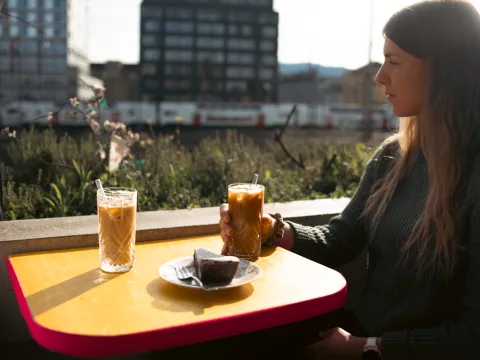 Une femme assise à une table buvant un café glacé. Elle regarde par-dessus son épaule droite en direction de la voie ferrée.