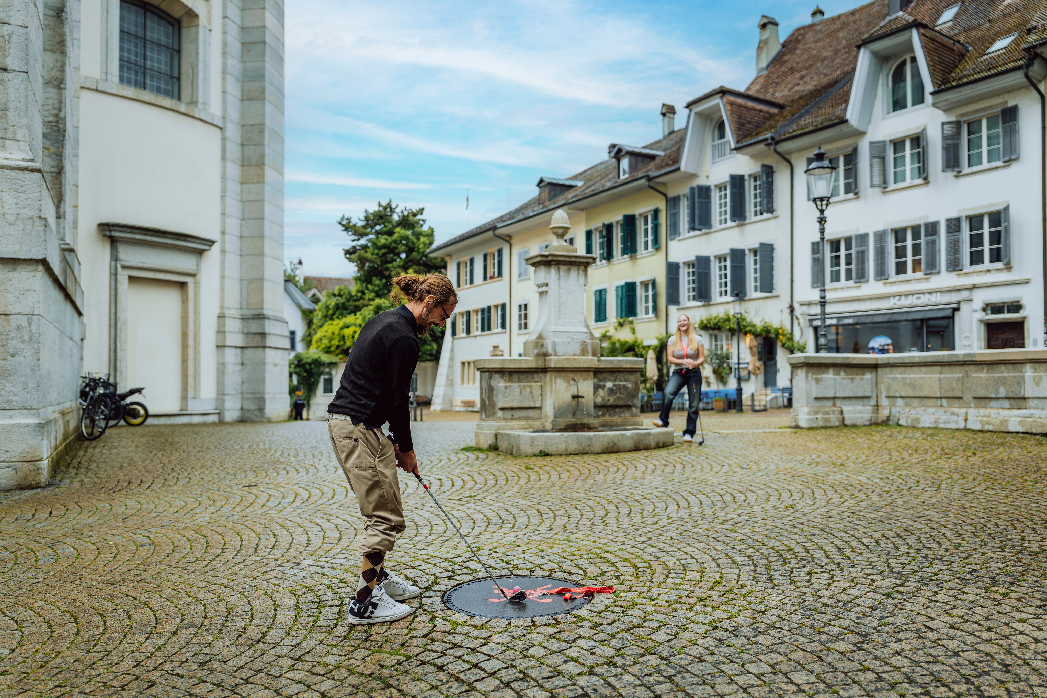 Eine Person mit einem Golfschläger auf einem Platz mit einem Brunnen, im Hintergrund Häuser und eine zweite Person mit einem Golfschläger.