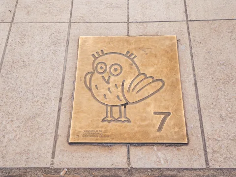 Una placca posizionata a terra, con una civetta e il numero sette incisi sul metallo.