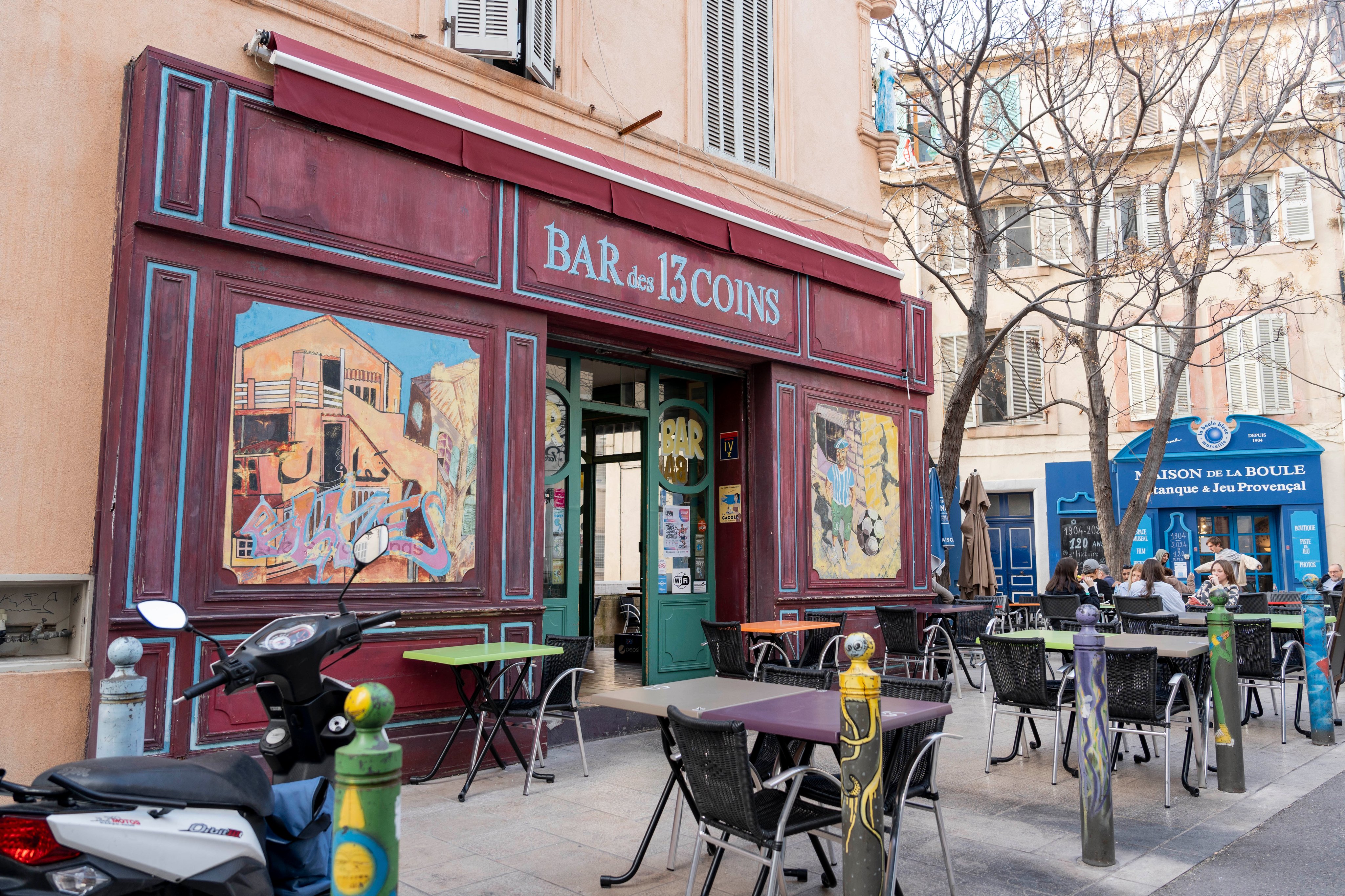 Vue extérieure du Bar des 13 coins à Marseille et de ses fresques murales colorées, au milieu de terrasses et d’arbres, par un jour ensoleillé.