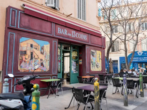 Vue extérieure du Bar des 13 coins à Marseille et de ses fresques murales colorées, au milieu de terrasses et d’arbres, par un jour ensoleillé.