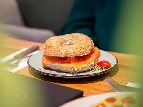 Auf einem Holztisch steht ein Teller, auf dem ein belegter Bagel liegt.