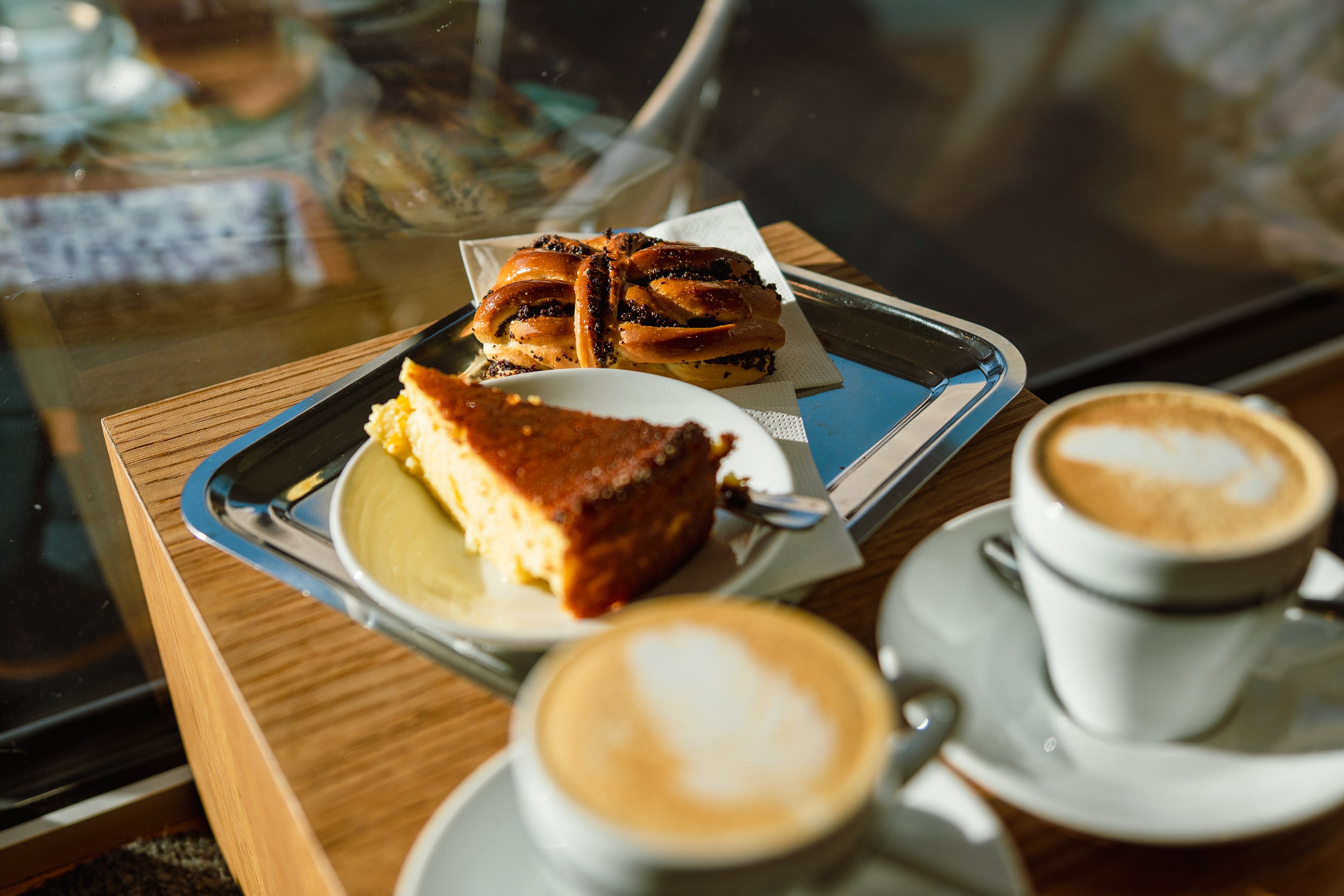 Su un piccolo tavolo di legno si trovano due caffè in tazze bianche con schiuma di latte e un vassoio argentato con una fetta di torta e un nodo di pan brioche.