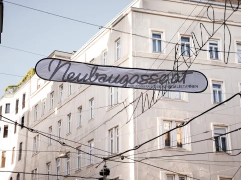 Uno striscione con la scritta «Neubaugasse» campeggia su una strada di Vienna. Intorno si vedono edifici di colore chiaro e le linee del tram.