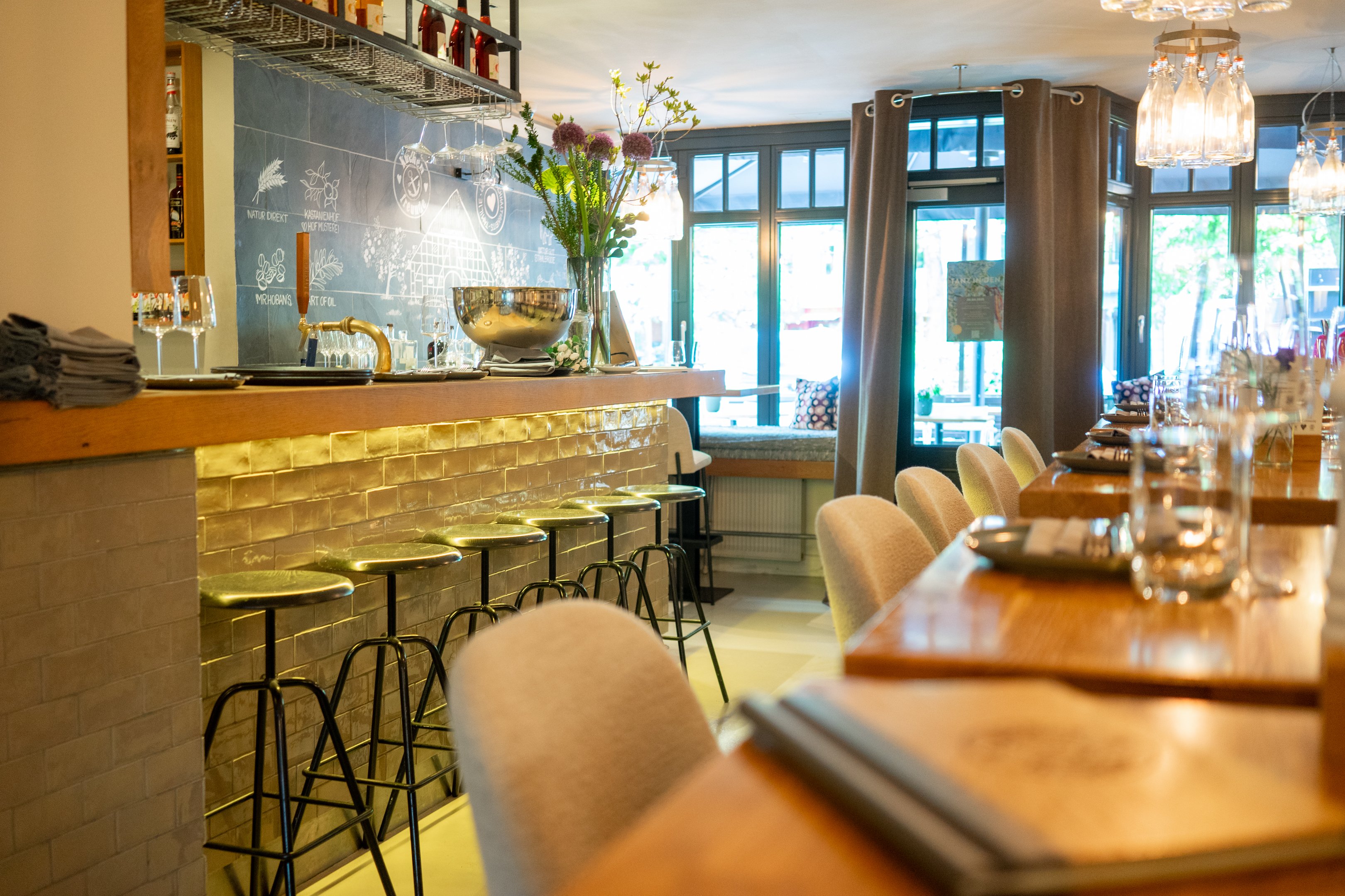 Lichtdurchfluteter Innenraum eines Restaurant mit einer Bar auf der Linken Seite und gedeckten Tischen auf der rechte Seite.