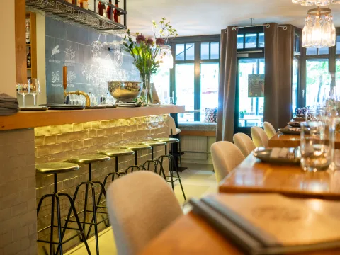 Lichtdurchfluteter Innenraum eines Restaurant mit einer Bar auf der Linken Seite und gedeckten Tischen auf der rechte Seite.