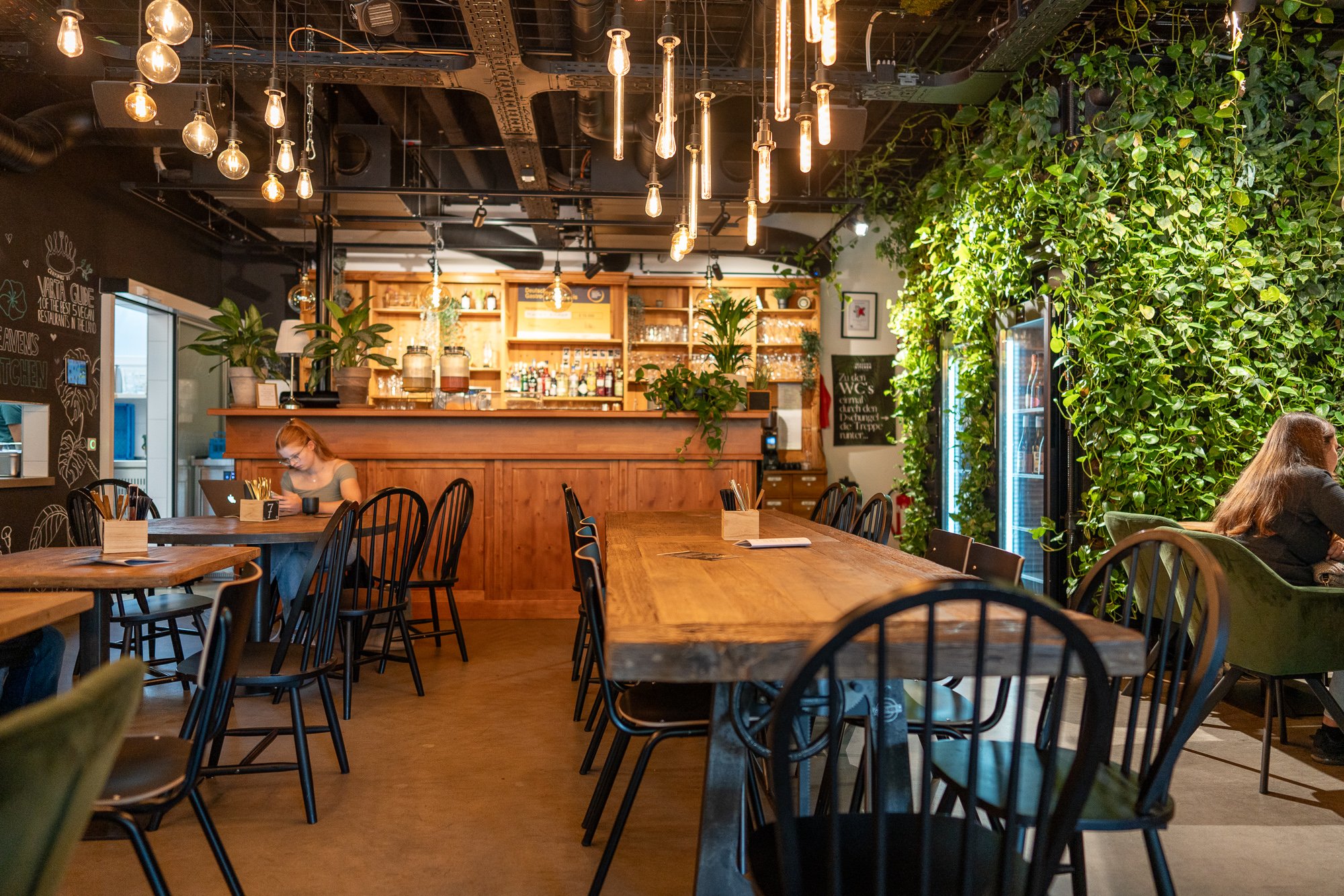 Un bar moderne, avec un comptoir en bois encombré de bouteilles et de verres. Au bar, vous pouvez prendre place, entre autres, sur une chaise en bois ou un fauteuil de velours vert. Les murs sont décorés d’objets culinaires et de plantes. Des lampes industrielles pendent au plafond. Le sol est en béton.