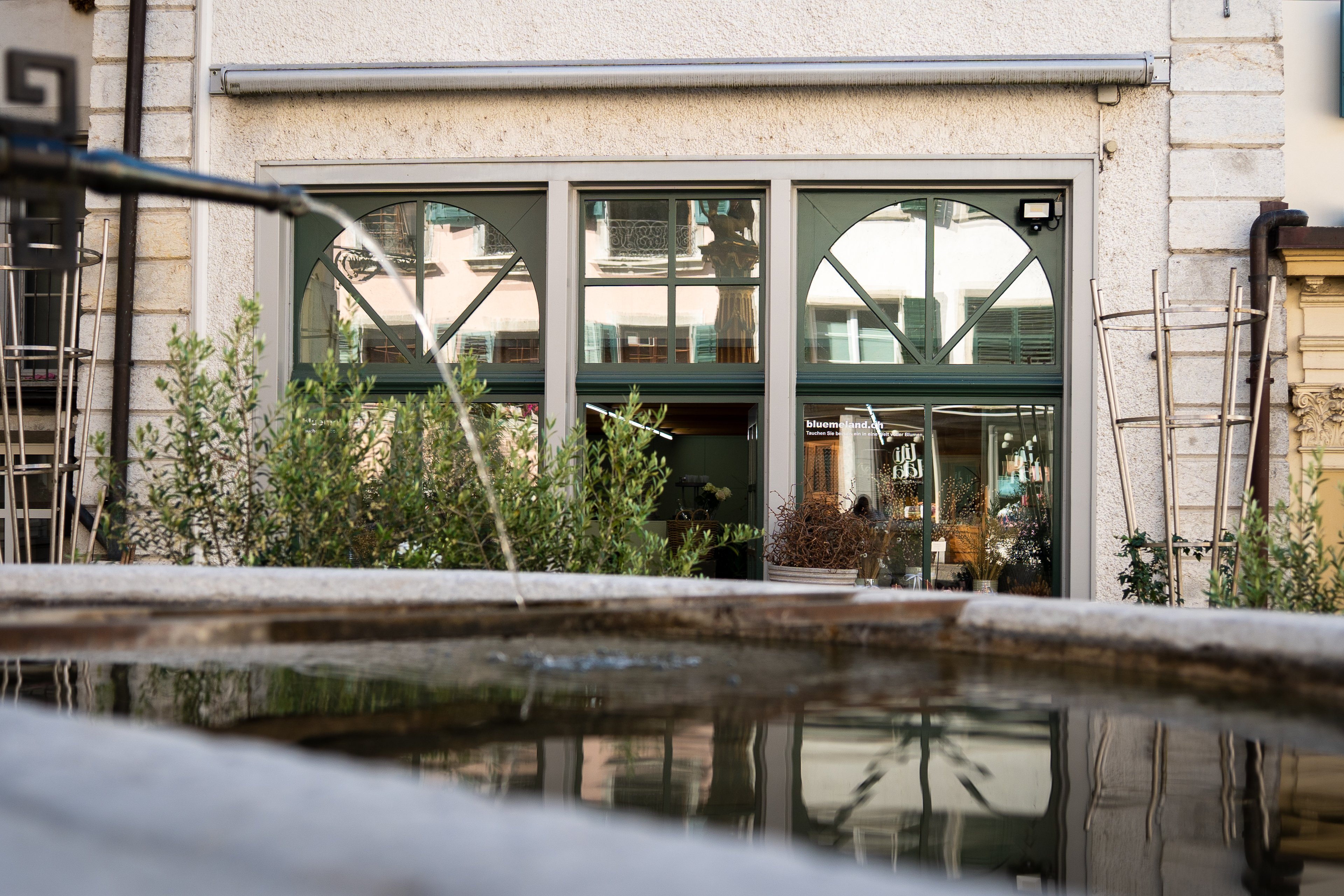 Im Vordergrund ein Brunnen, im Hintergrund ein Gebäude mit hohen Fenstern.