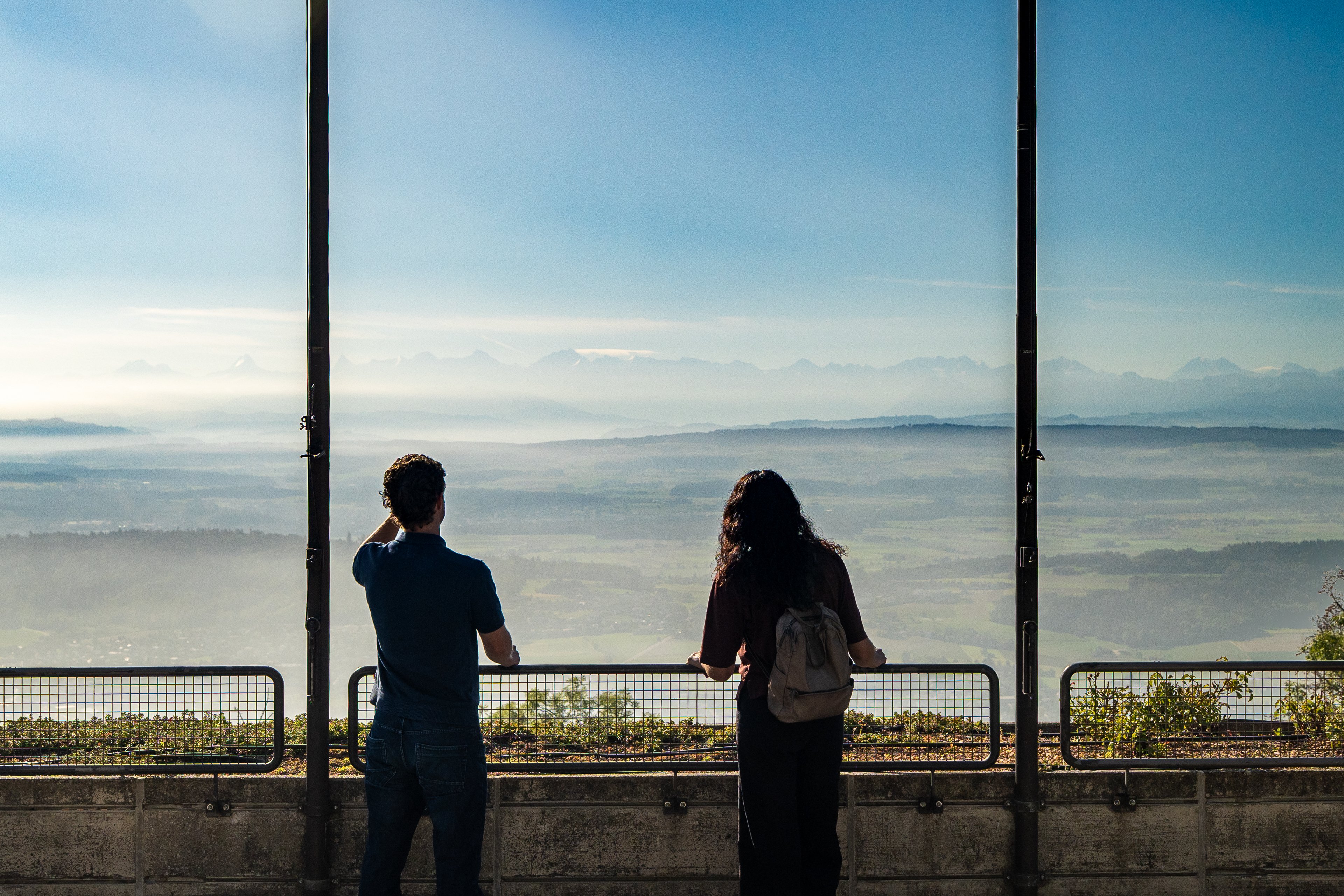 Due persone sono appoggiate alla ringhiera della piattaforma panoramica di Macolin. Sullo sfondo si distinguono verdi colline e la catena alpina.