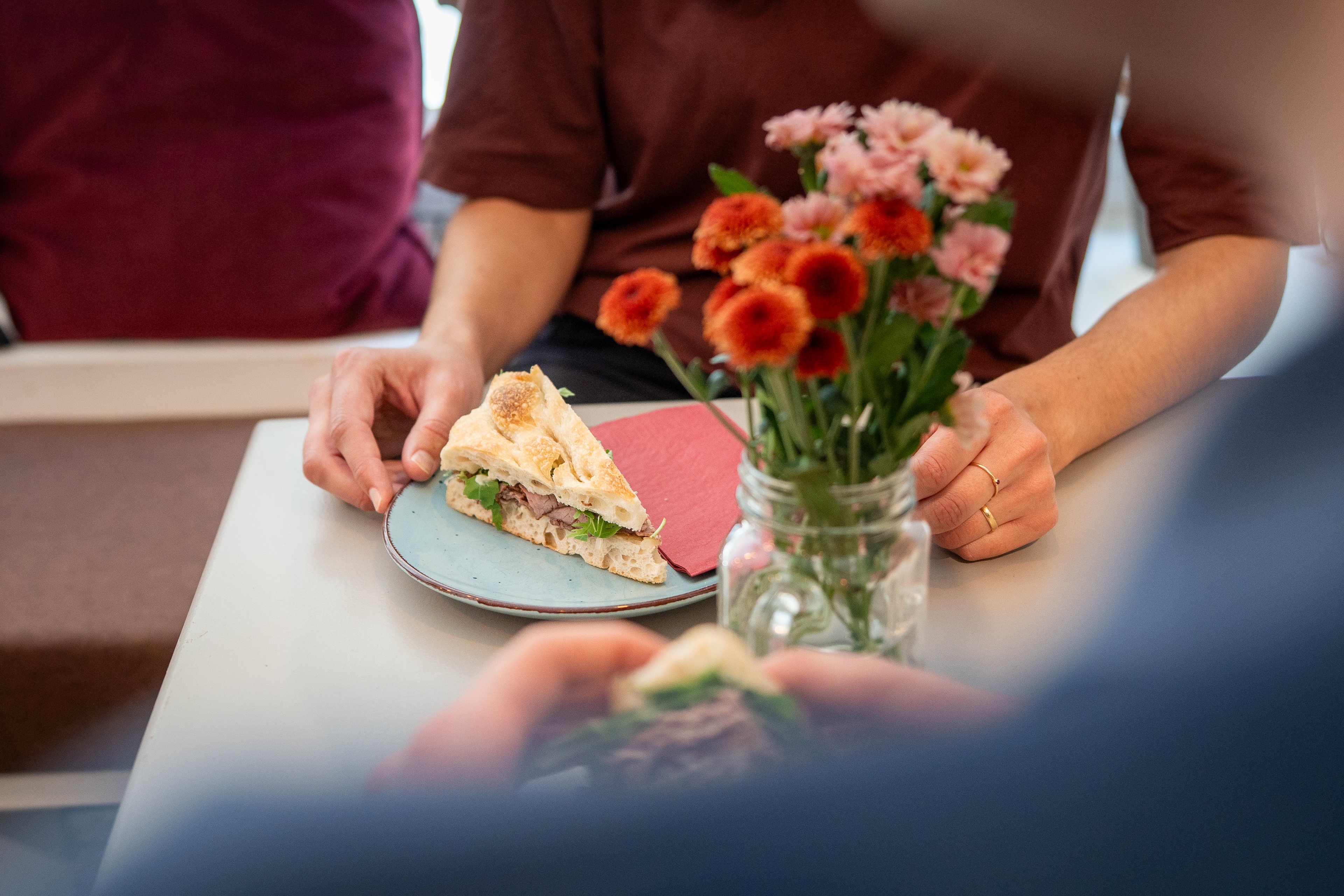 Su un tavolo si trova un variopinto mazzo di fiori in un vaso di vetro. Si vede, inoltre, un piattino con un tovagliolo e un sandwich.