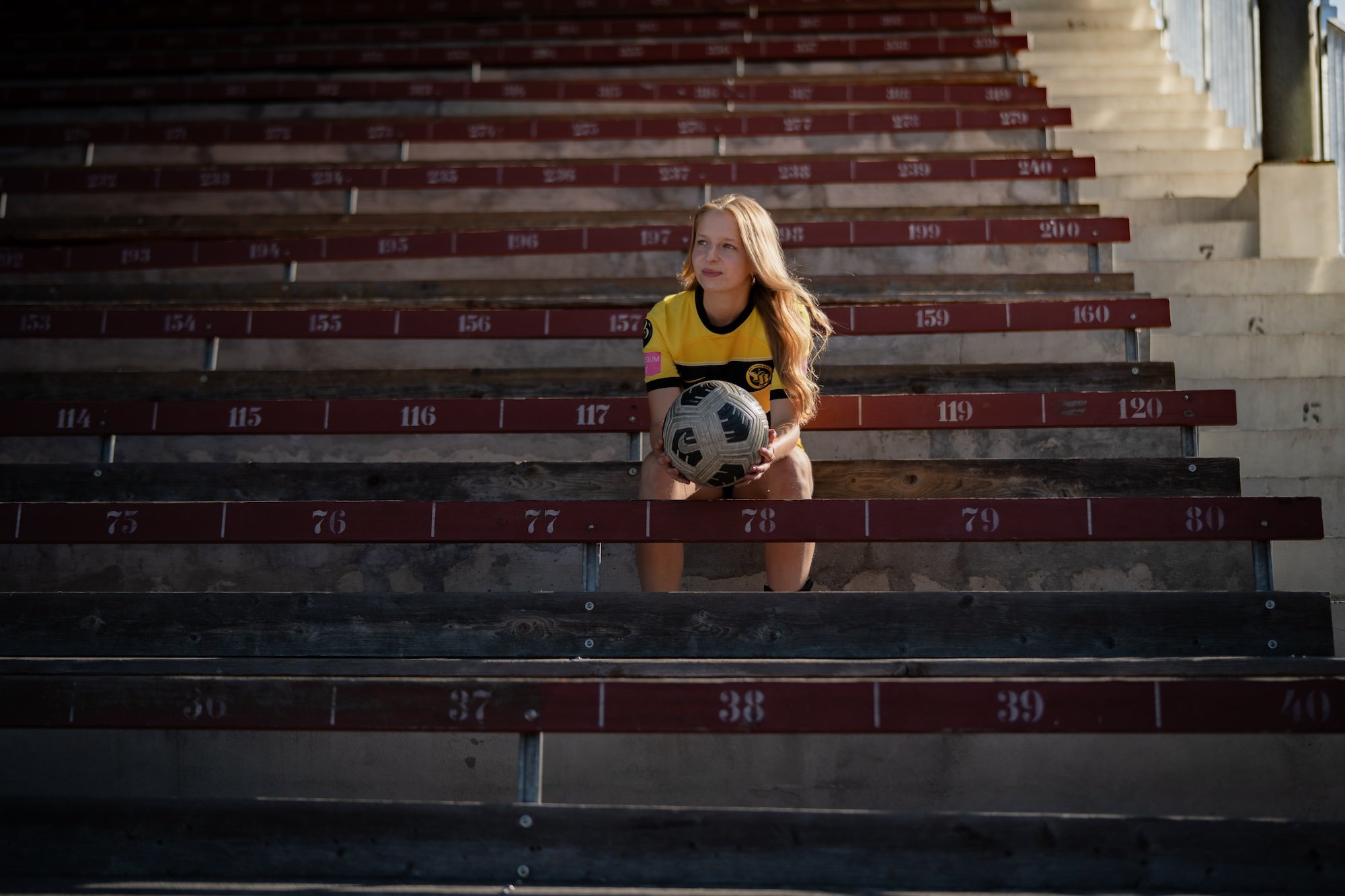 Una donna in tenuta sportiva siede in tribuna con lo sguardo trasognato e una palla in mano.