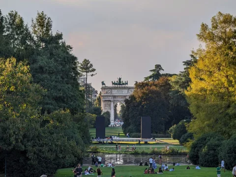 Delle persone sono sdraiate e sedute sull’erba del parco. Sullo sfondo si vede una costruzione a forma di arco.