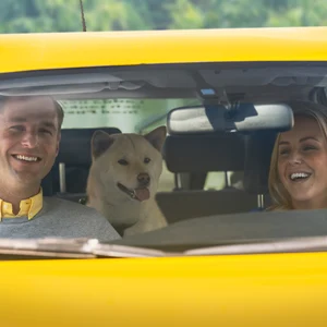 Två personer sitter och skrattar i en gul bil. En hund tittar fram från baksätet.