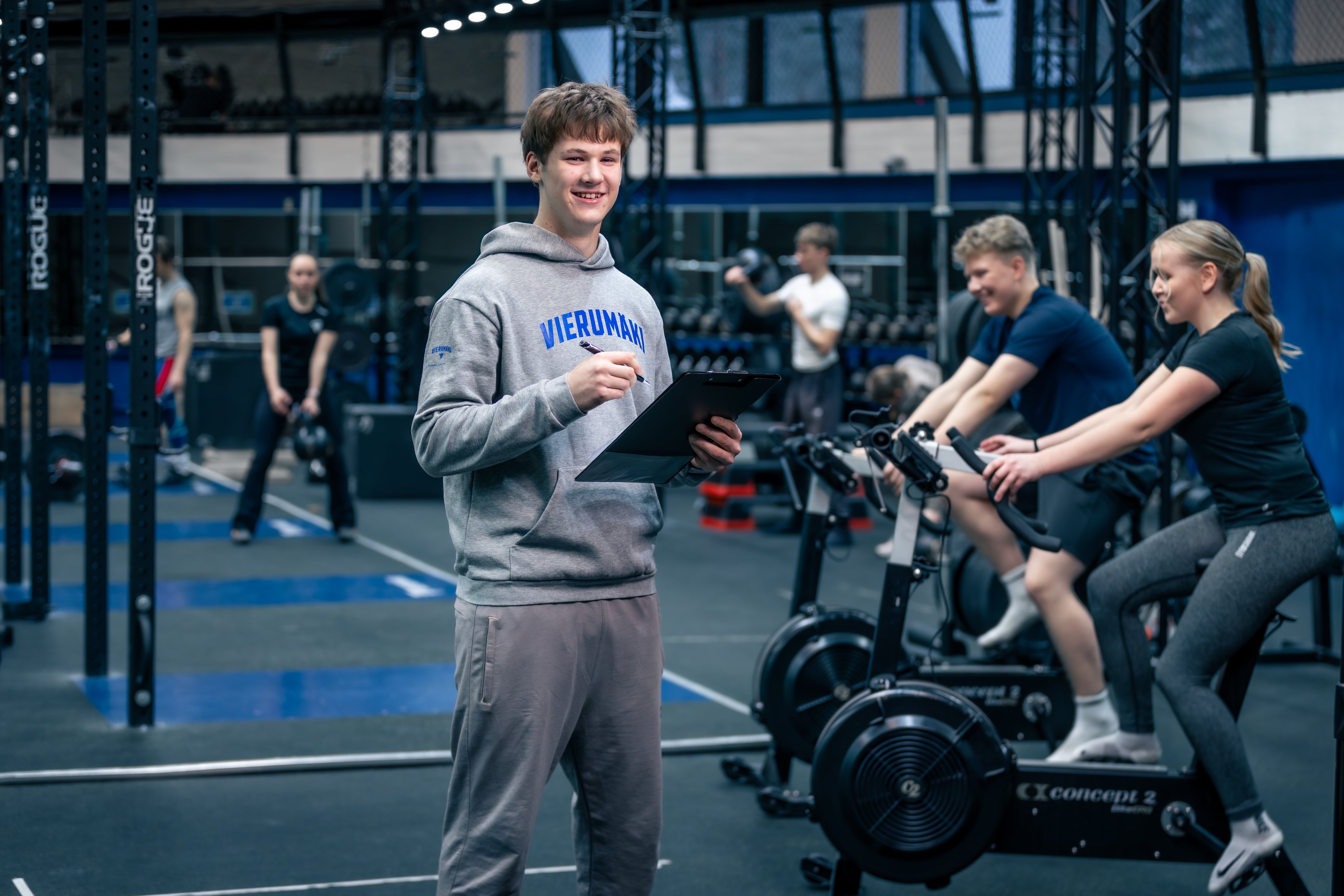 Ohjaaja kuntosalilla. Kaksi ihmistä pyöräilee vieressä.