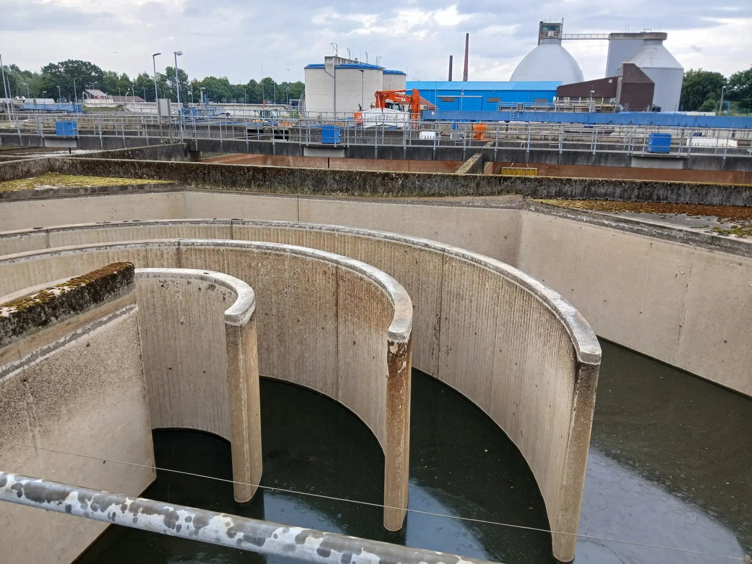 Wasserrückhaltebecken mit vertikalen Betonwänden und Wasser im unteren Bereich, im Hintergrund Gebäude