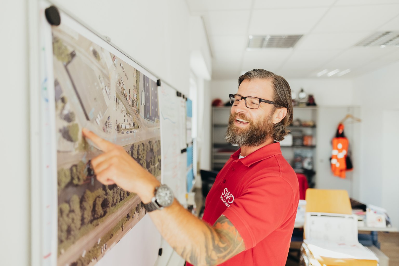 Mann mit Brille und Tätowierung zeigt auf eine Karte an einer Wand im Büro