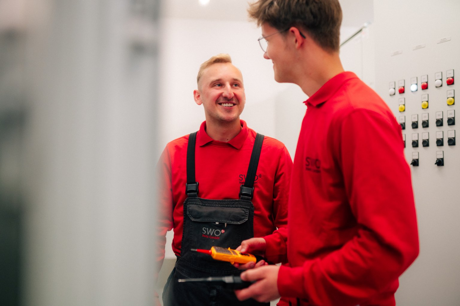 Zwei männliche Personen in rot-gestreiften Oberteilen sprechen in einem Technikraum.