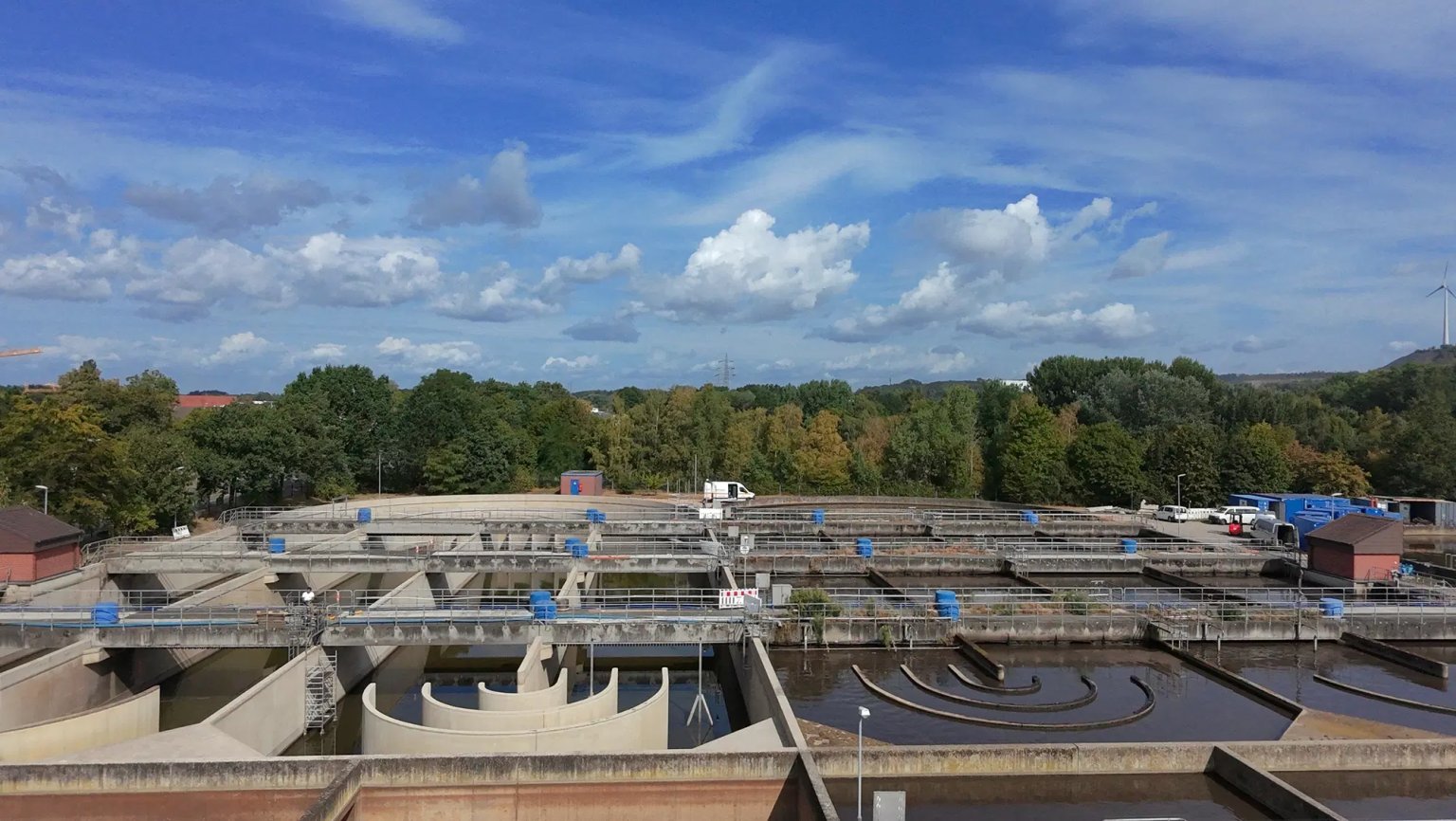 Blick auf eine Kläranlage mit Becken, Wasser und Baufahrzeugen, umgeben von Bäumen und Himmel
