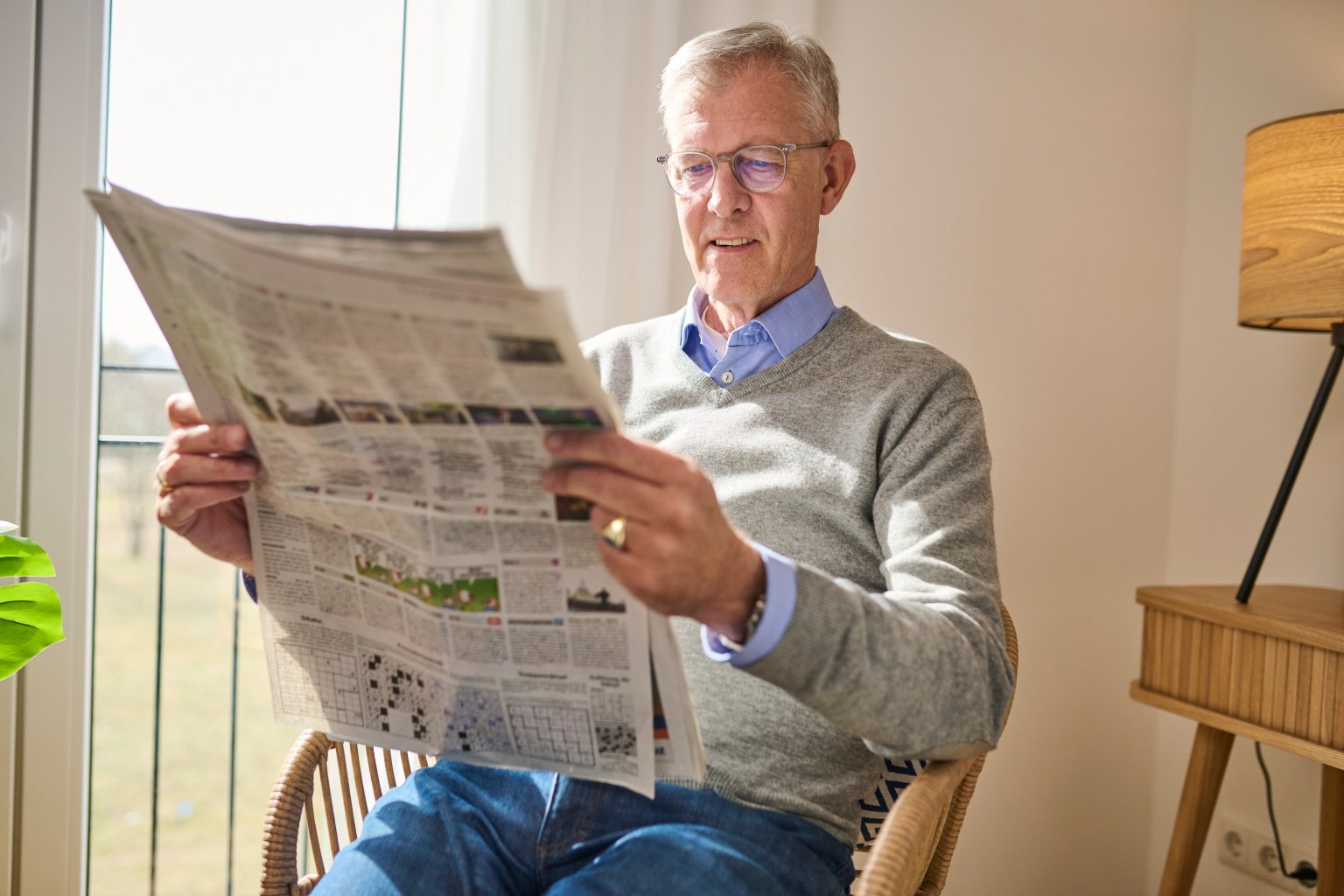 Älterer Mann liest Zeitung in hell erleuchtetem Raum, neben Stehlampe.