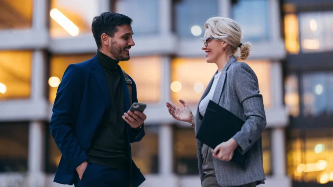 A man and a woman share a friendly conversation outside a modern business building, smiling warmly