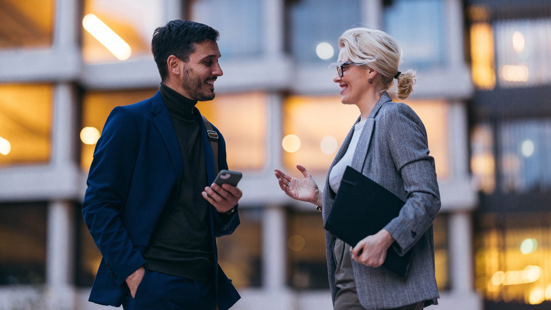 A man and a woman share a friendly conversation outside a modern business building, smiling warmly
