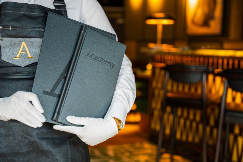 Academy Restaurant team member holding the restaurant menu in a contemporary dining room.