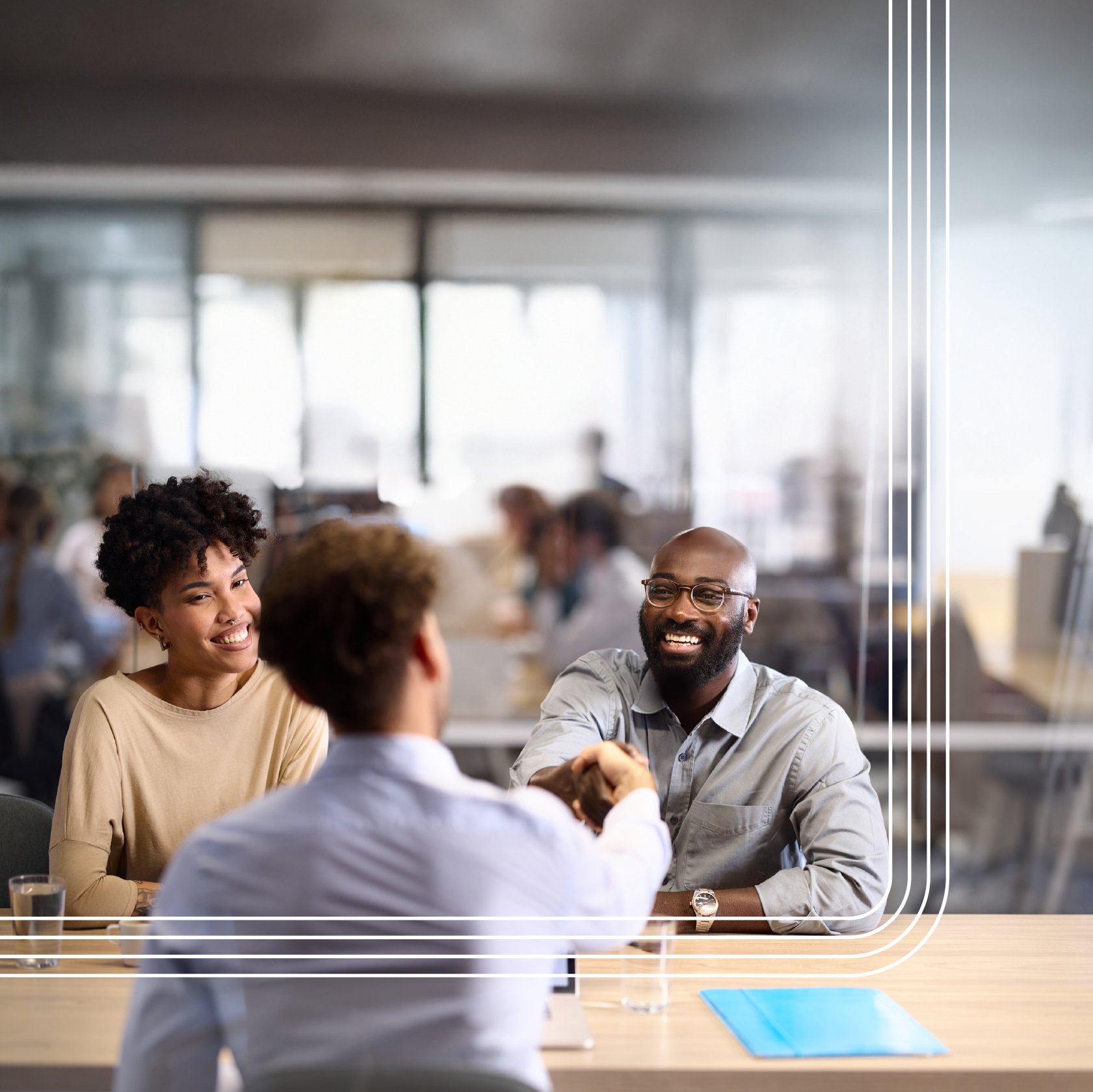 A smiling man and woman sit at a table in a modern office, while the man shakes hands with another person across from them.