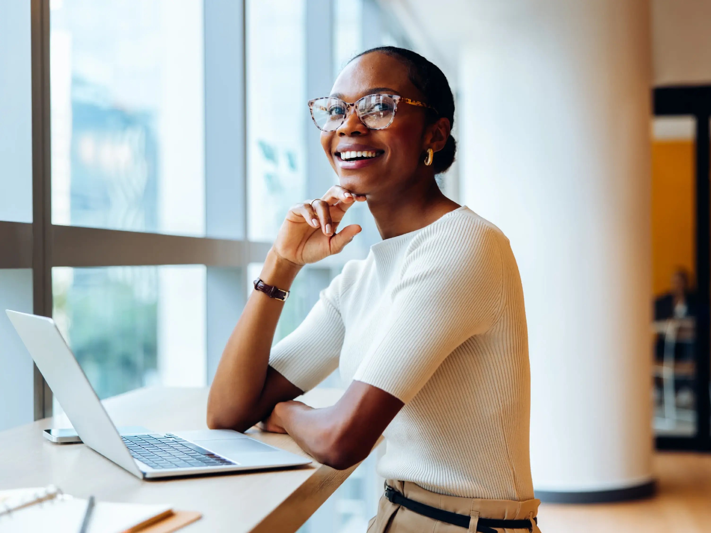 Professional woman smiling while working on a laptop by a window in a modern office setting, wearing glasses and a white top