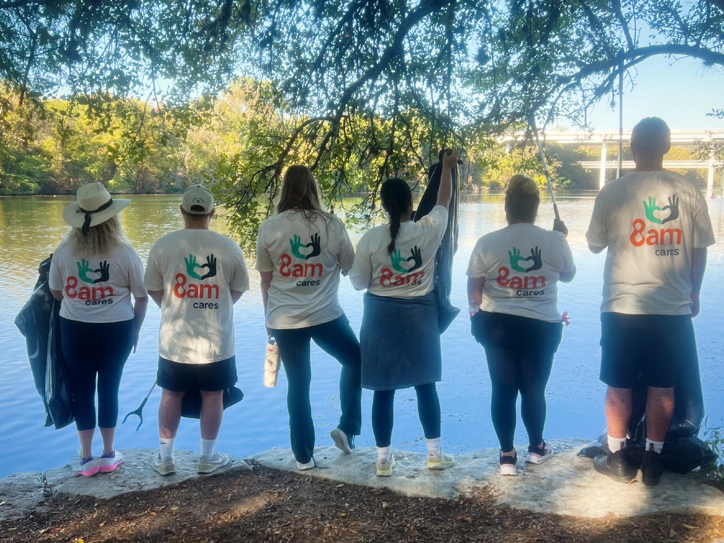 Six volunteers wearing white ‘8am cares’ shirts stand along a riverbank holding trash bags and cleanup tools during a community river cleanup.
