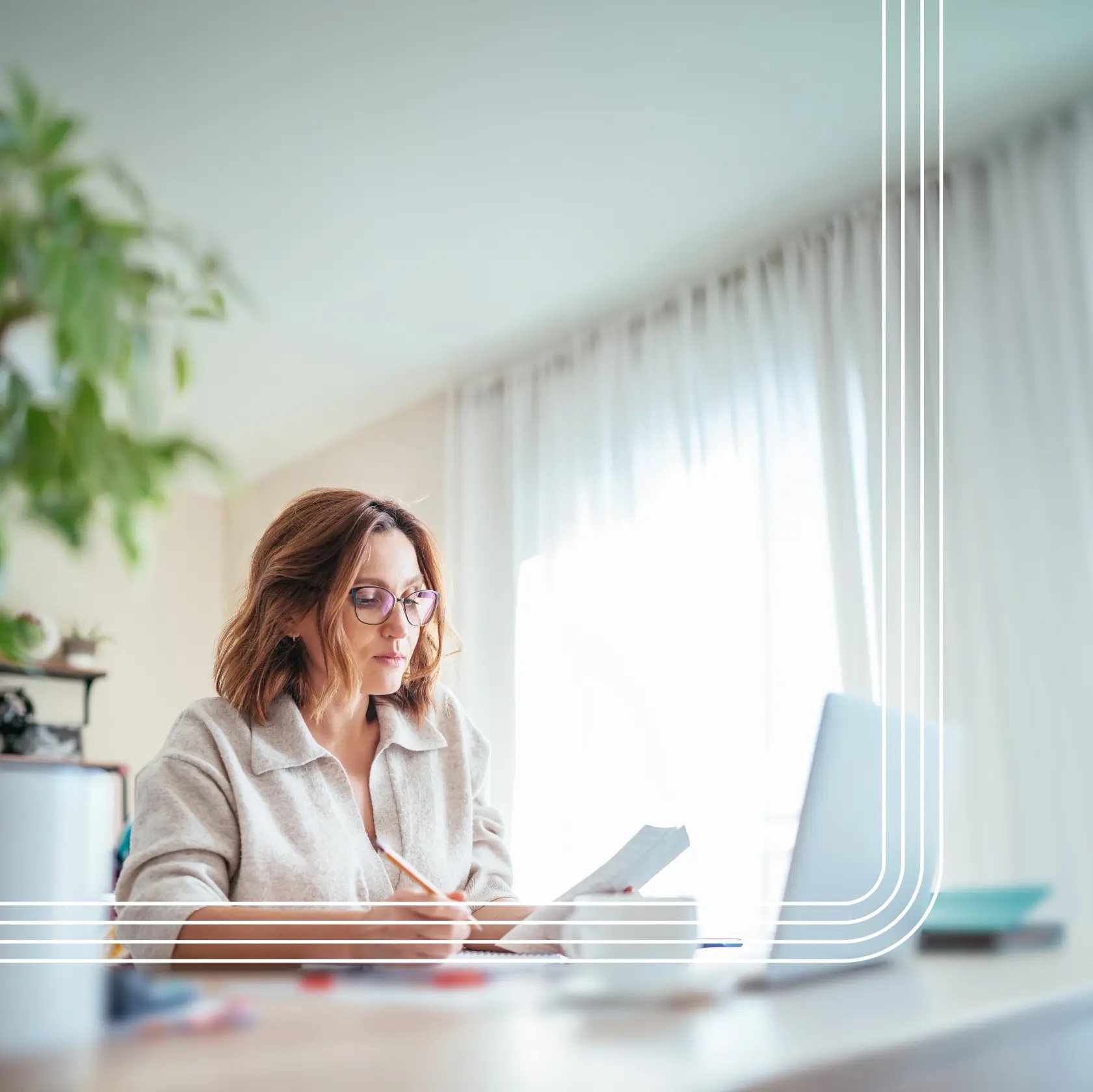 Woman wearing glasses sits at a desk, reviewing papers and writing notes beside an open laptop in a bright, home office setting.