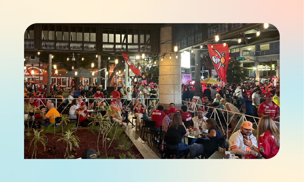 Crowded outdoor patio scene at the Tampa Bay Buccaneers Draft Block Party, with fans gathered under string lights and Buccaneers flags in downtown St. Petersburg.
