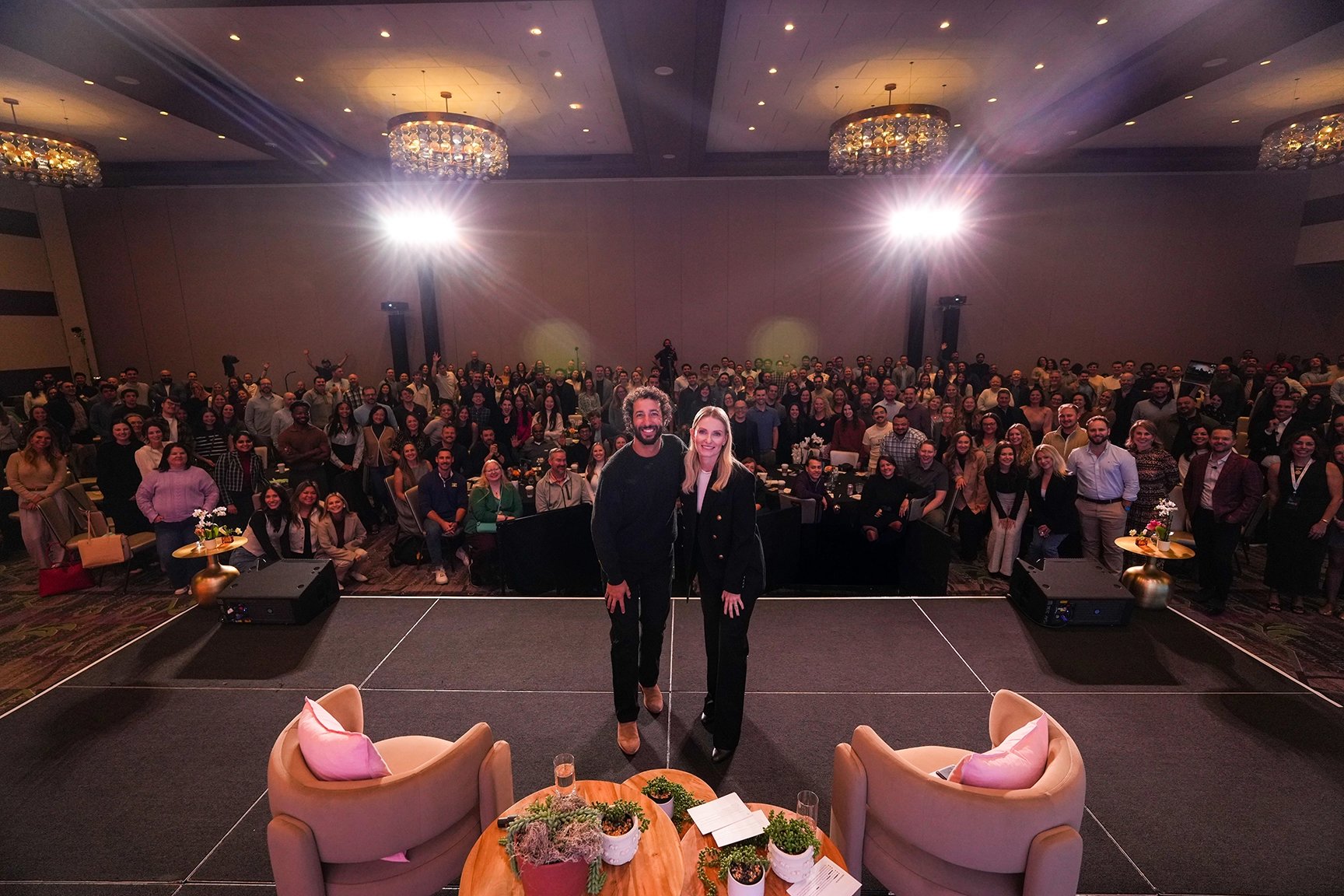 8am CEO Dru Armstrong and F1 Driver Daniel Ricciardo pose in front of a large crowd of 8am employees after their talk during the 2026 Company Kick Off