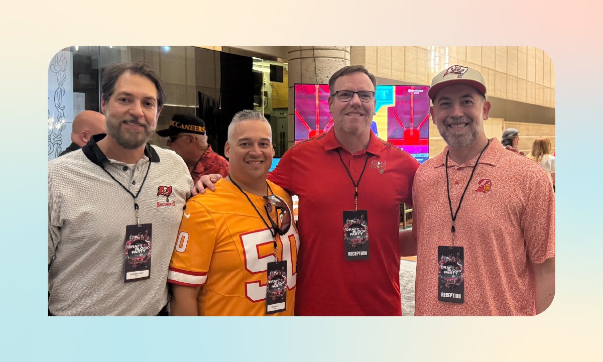 Four people wearing Buccaneers apparel and event credentials pose together inside the Tampa Bay Buccaneers Draft Block Party reception area.