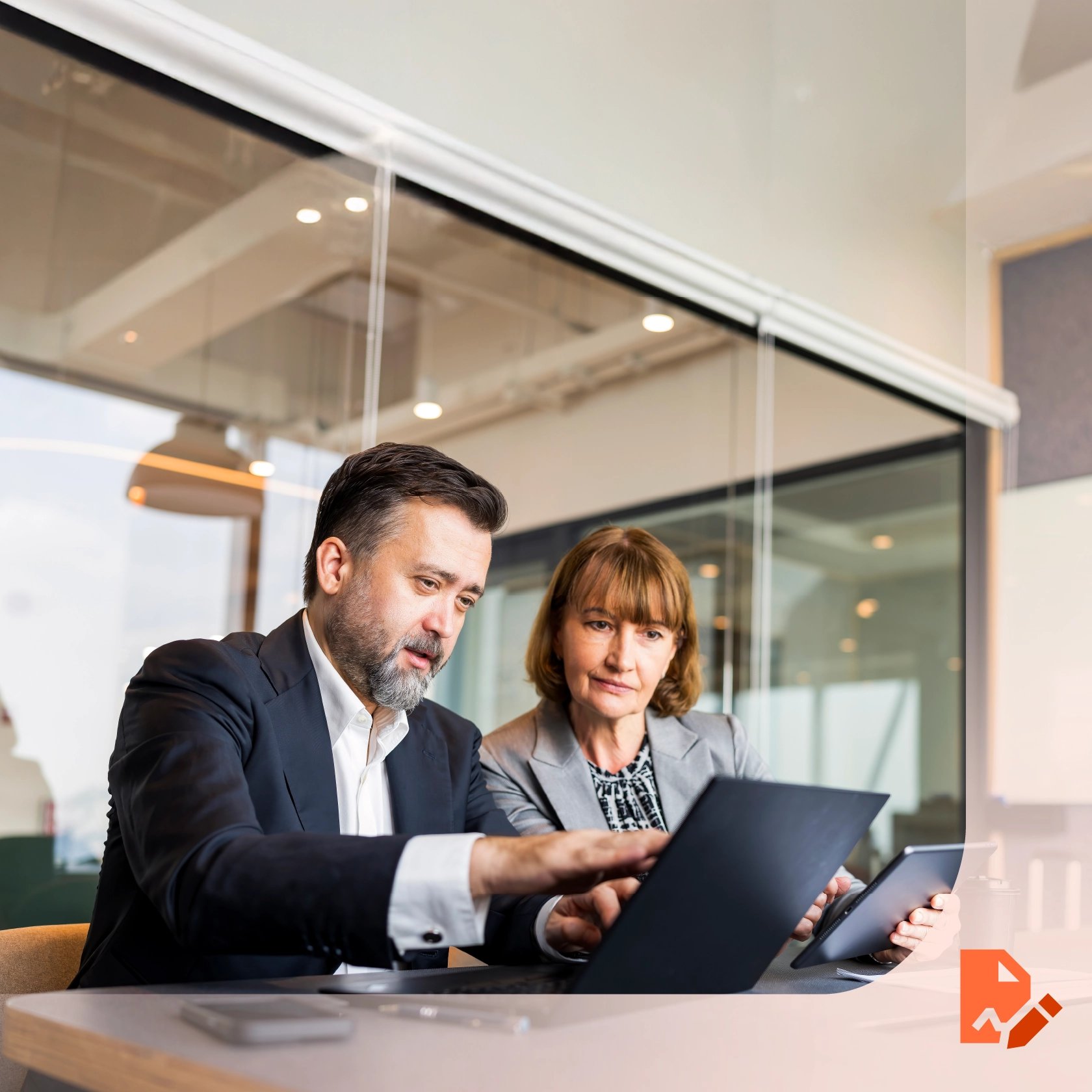 Two business professionals sitting at a table in office, looking at a laptop screen together. Large windows and glass walls are visible in the background.