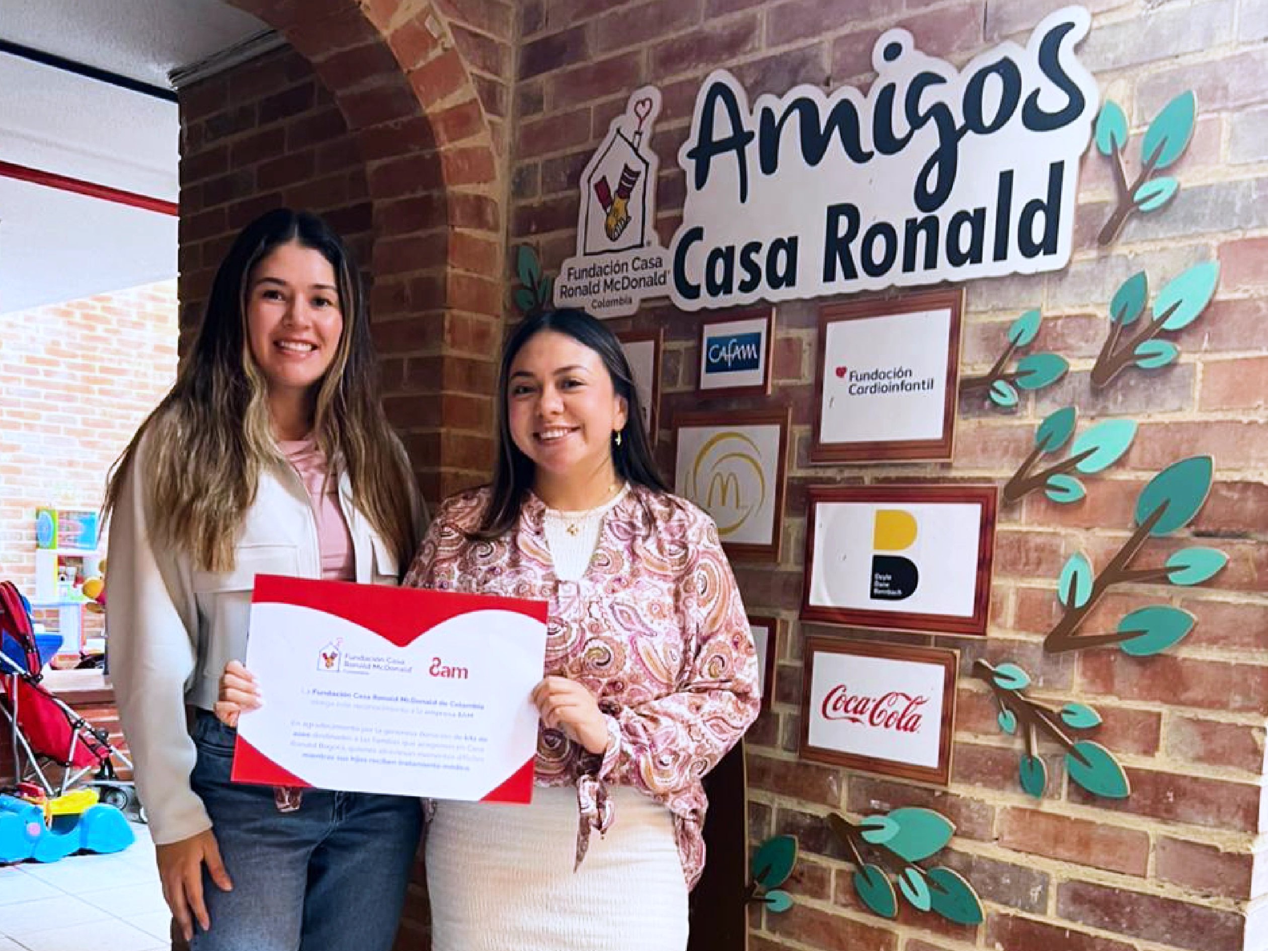 Two women stand in front of an ‘Amigos Casa Ronald’ wall holding a certificate from 8am, representing a partnership with Fundación Casa Ronald McDonald Colombia.