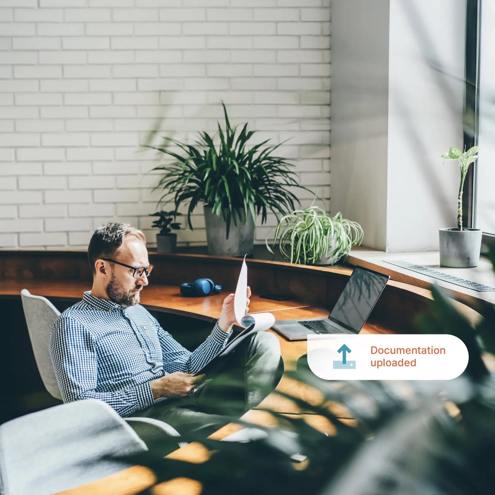 A man sits in a modern, bright room with plants, reading papers and using a laptop on his lap. An arrow and text overlay say “Documentation uploaded"