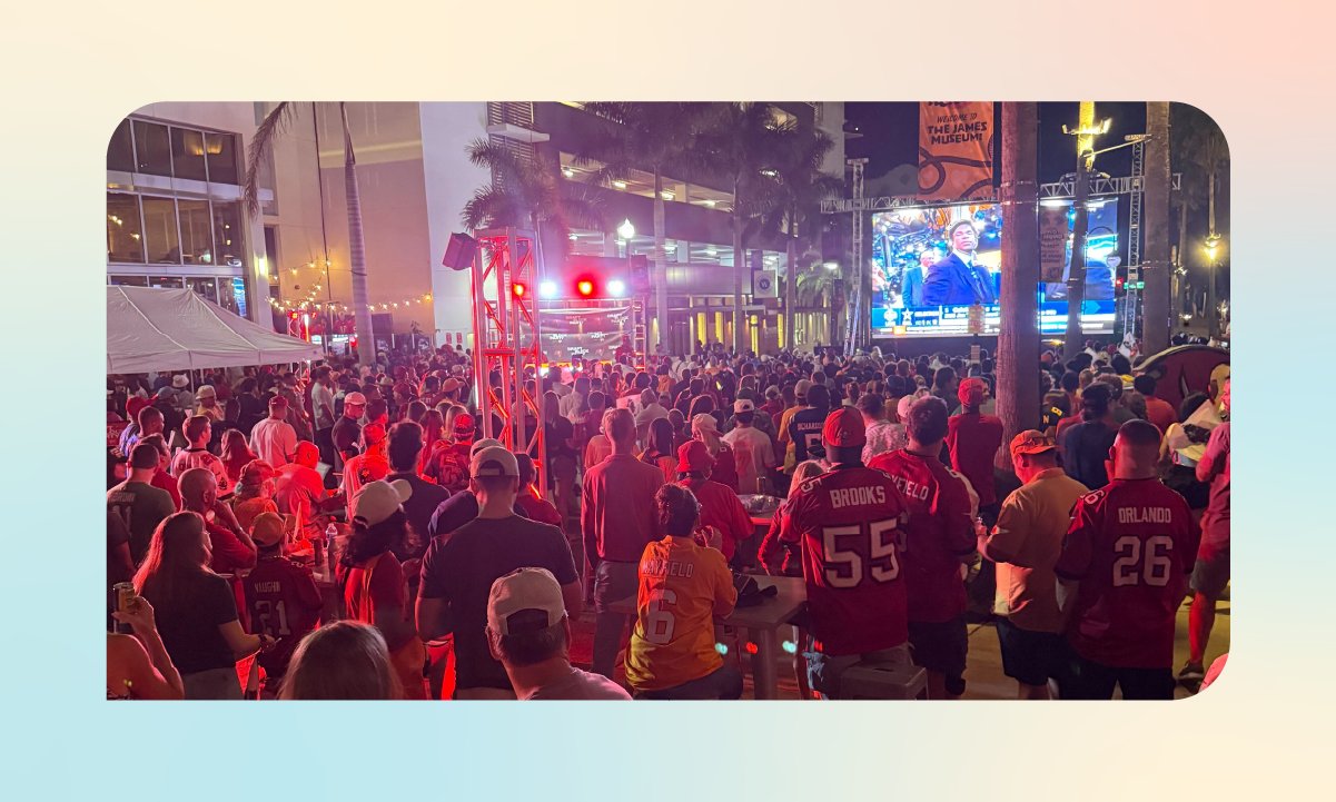 Large crowd watching a broadcast on an outdoor screen during the Tampa Bay Buccaneers Draft Block Party at night.