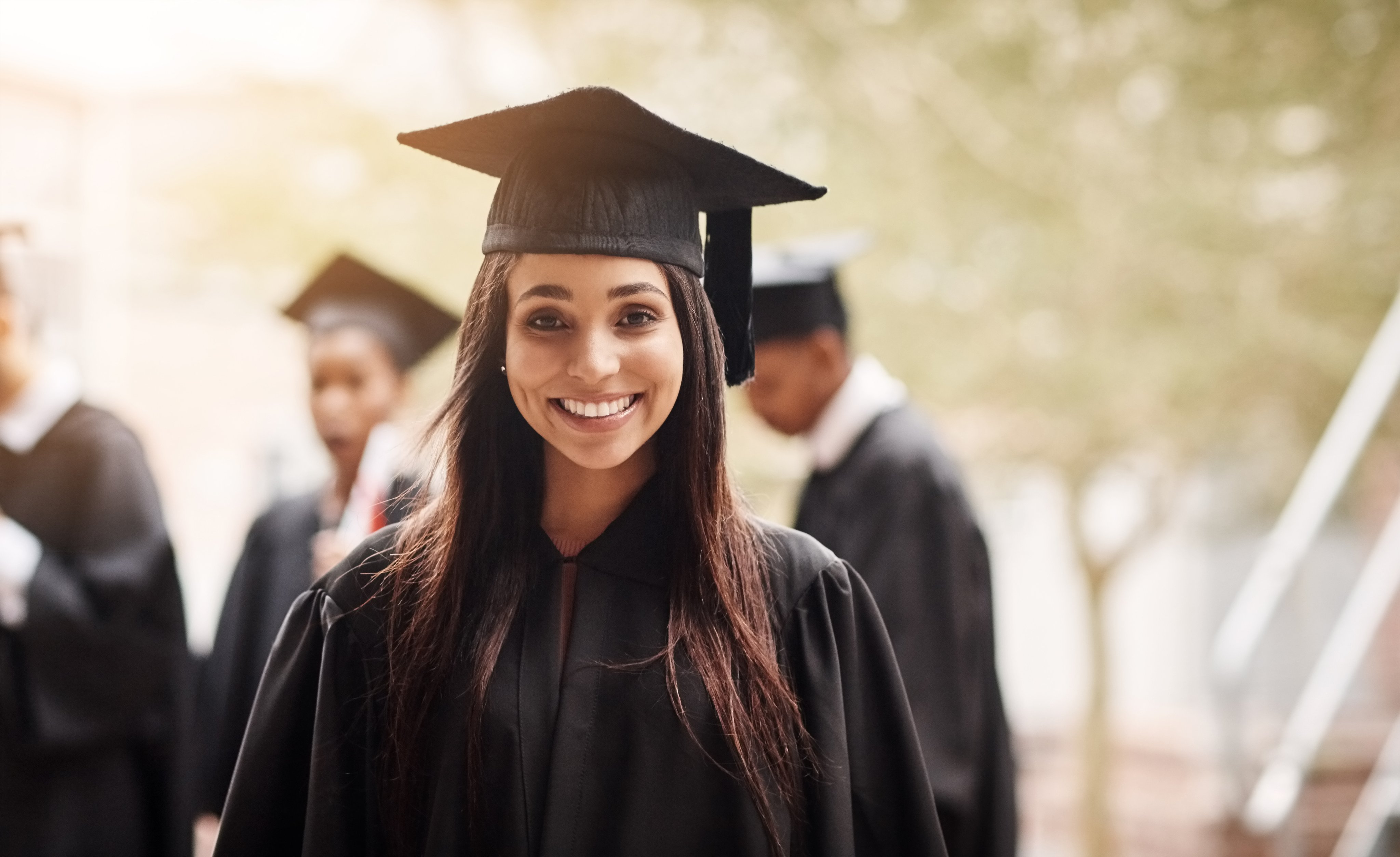 Portrait of a smiling female graduate in cap and gown, with fellow graduates standing in the background.