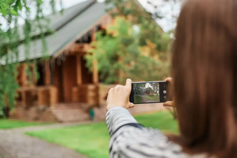 Back view of brown haired woman photographing wooden house for insurance reasons